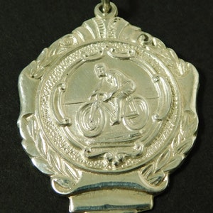 May include: Silver medal with a cyclist riding a bicycle in the center. The medal is surrounded by a laurel wreath and has a loop for hanging.