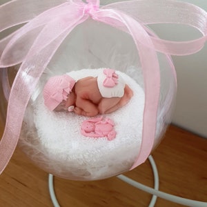 Op de afbeelding: Een transparante glazen ornament met een roze lint en een kleine babypop erin. De babypop draagt een roze muts en een witte luier met een roze strik. De ornament staat op een witte standaard.
