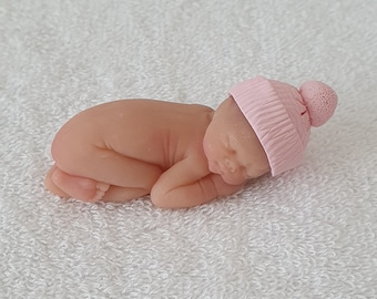 May include: A small, sleeping baby doll made of a soft, flesh-toned material. The doll is wearing a pink knitted hat with a white pom-pom.