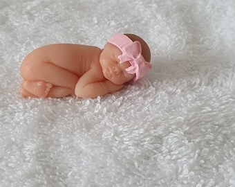 May include: A small, sleeping baby doll with a pink bow headband. The doll is lying on a white, fluffy towel.