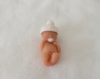 May include: A small, white ceramic baby doll wearing a white knit hat and a pacifier. The doll is lying on its back with its arms and legs slightly bent.