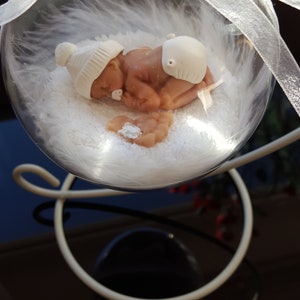 May include: A glass bauble with a white feathery interior, containing a small, sleeping baby doll wearing a white hat and nappy. The bauble is suspended from a white ribbon and sits on a white stand.