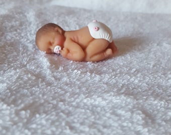 May include: A small, sleeping baby doll made of fondant, wearing a white diaper with pink polka dots.