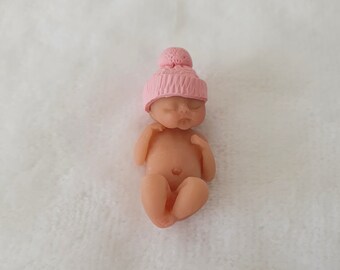 May include: A small, realistic baby doll with a pink knitted hat. The doll is lying on its back with its eyes closed.