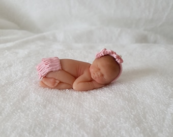 May include: A small, sleeping baby doll made of fondant, wearing a pink ruffled diaper and a pink headband with white accents.