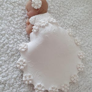 Könnte beinhalten: Schlafende Babyfigur mit weißem Blumenstirnband und weißem Kleid mit Blumen verziert. Das Kleid hat einen gewellten Rand und florale Details. Die Figur liegt auf einer weißen, strukturierten Oberfläche.