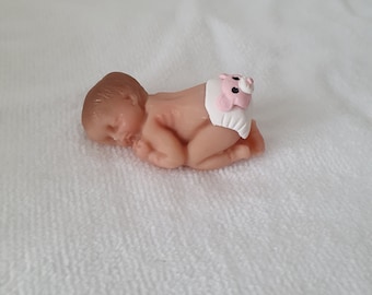 May include: A small, sleeping baby doll made of painted resin. The doll is wearing a white diaper with a pink bear design.