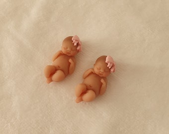 May include: Two small, sleeping baby dolls with pink bows on their heads. The dolls are made of a soft, realistic material and are lying on a white, fluffy surface.
