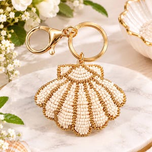 May include: A seashell-shaped keychain featuring intricate beadwork in cream and metallic gold. The design consists of rows of small cream-colored beads separated by lines of tiny gold beads. It is attached to a polished gold-tone metal keyring.