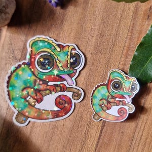 May include: Two colourful chameleon stickers with a cartoon design. The stickers are green, red, and yellow with glitter accents. One sticker is larger than the other. The stickers are on a wooden surface with purple flowers.