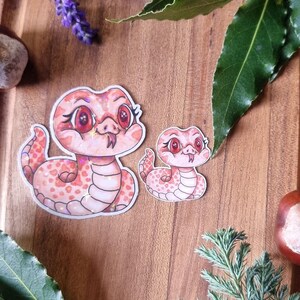 May include: Two pink snake stickers with red eyes and a white belly are displayed on a wooden surface. The stickers are surrounded by sprigs of lavender, green leaves, and a chestnut. The stickers have a cartoonish design.