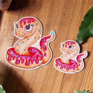 May include: Two stickers featuring a cartoon snake design. The snakes are shaped like the number 2, with a pink frosting and sprinkles design. The stickers are on a wooden surface with lavender and greenery.