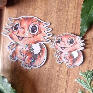 May include: Two orange and red bearded dragon stickers with glitter details. The stickers have large eyes and are smiling. They are on a wooden surface with purple flowers and green leaves.