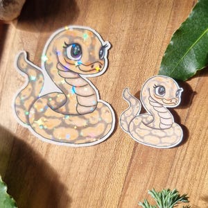 May include: Two cartoon snake stickers with glitter detailing. The snakes have tan and brown patterns and large eyes. They are set on a wooden surface, with sprigs of lavender and greenery in the background.