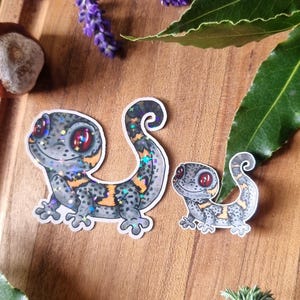 May include: Two gecko stickers with grey, orange, and iridescent details are displayed on a wooden surface. Lavender sprigs, a chestnut, and green leaves surround the stickers. The gecko stickers have red eyes and curled tails.