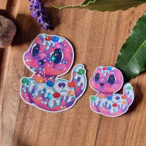 May include: Two colourful stickers of a cartoon creature with a pink and blue colour scheme, decorated with sprinkles and glitter. The stickers are on a wooden surface, with lavender and greenery in the background.