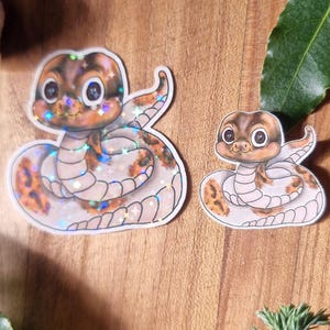 May include: Two cartoon snake stickers. The larger one has a coiled design with brown and orange markings, and the smaller one has a similar pattern. Both have a holographic finish and are on a wooden surface.