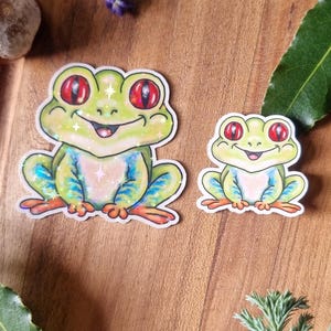May include: Two frog stickers with red eyes and green and blue bodies are displayed on a wooden surface. Lavender and greenery are arranged around the stickers. The stickers have a whimsical, cartoonish design.