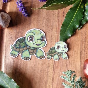 May include: Two green turtle stickers with large eyes and detailed shells are arranged on a wooden surface, accompanied by lavender sprigs, green leaves, and conkers. The stickers are of different sizes, with the larger one in the foreground.