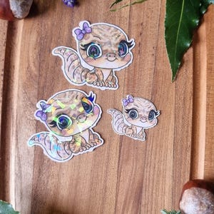 May include: Three cartoon lizard stickers with iridescent details and purple bows are displayed on a wooden board. Lavender, green leaves, and conkers surround the stickers, creating a whimsical, natural scene.
