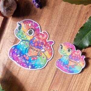 May include: Two colourful stickers featuring a cartoon snake holding an ice cream cone. The stickers have a rainbow colour scheme with hearts and stars. The stickers are on a wooden surface with lavender and greenery.