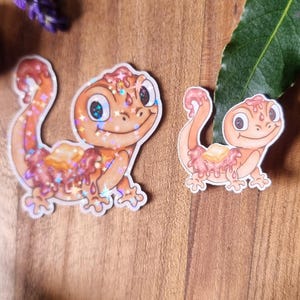 May include: Two stickers of a cartoon gecko in orange and pink. The gecko has a smiling face and a glittery, textured design. The stickers are on a wooden surface with lavender and greenery.