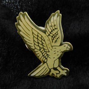 Könnte beinhalten: Eine goldfarbene Anstecknadel mit einer weißen Umrandung eines fliegenden Vogels. Der Vogel hat detaillierte Federn und einen Schnabel.