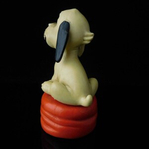 May include: A white ceramic figurine of a dog sitting on a red and orange cushion. The dog has black ears and a black nose.