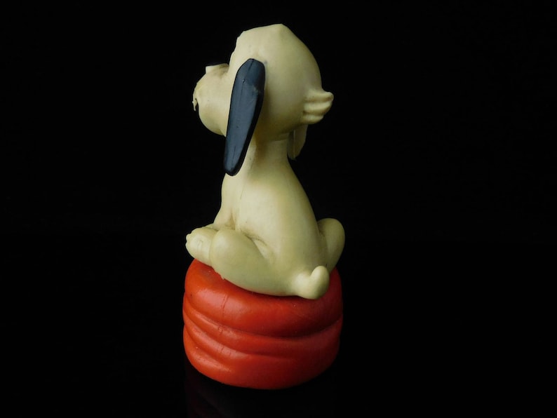 May include: A white ceramic figurine of a dog sitting on a red and orange cushion. The dog has black ears and a black nose.