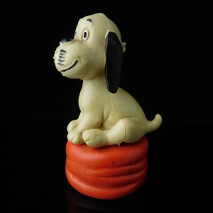 May include: A beige and black plastic toy dog sitting on a red and orange plastic base. The dog has a black nose and black ears.