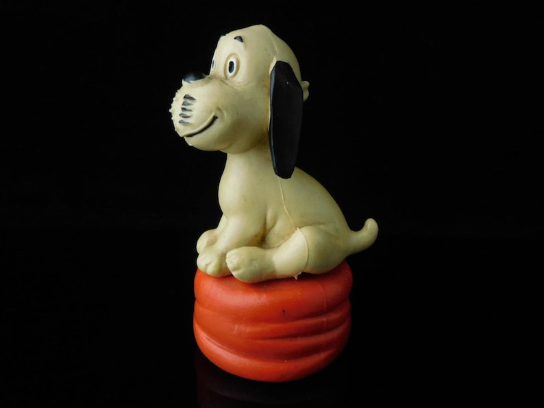 May include: A beige and black plastic toy dog sitting on a red and orange plastic base. The dog has a black nose and black ears.