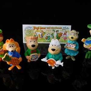 Peut inclure: Un ensemble de sept figurines de Yogi l'ours et ses amis. Les figurines sont en plastique et sont de couleurs vives. Elles comprennent Yogi l'ours, Boo Boo, Cindy l'ourse, Ranger Smith et trois autres personnages. Les figurines sont toutes debout sur un fond noir.