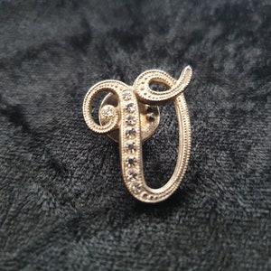 May include: A silver-toned letter V brooch with clear rhinestones. The letter is in a cursive font and the rhinestones are set in a line around the edge of the letter.