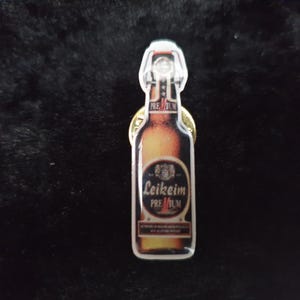 Könnte beinhalten: Ein goldfarbener Flaschenöffner in Form einer Bierflasche mit einem Etikett, auf dem "Leikeim Premium" steht.