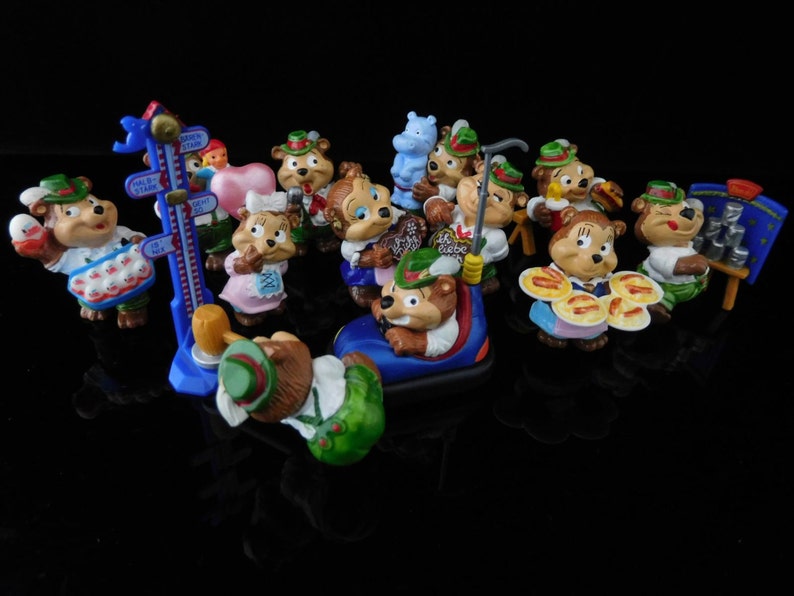 May include: A collection of 14 small, colorful figurines of bears wearing traditional German clothing. The figurines are made of plastic and have a variety of poses and expressions. Some of the bears are holding food, such as pretzels and sausages, while others are holding musical instruments. The figurines are all standing on a black surface.