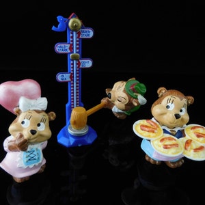 May include: Three small squirrel figurines with different outfits. One is wearing a pink dress and a heart-shaped hat, another is wearing a green hat and a brown vest, and the third is wearing a blue dress and holding a plate of sausages. The figurines are standing in front of a blue and white measuring stick with German text.