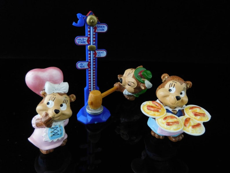 May include: Three small squirrel figurines with different outfits. One is wearing a pink dress and a heart-shaped hat, another is wearing a green hat and a brown vest, and the third is wearing a blue dress and holding a plate of sausages. The figurines are standing in front of a blue and white measuring stick with German text.