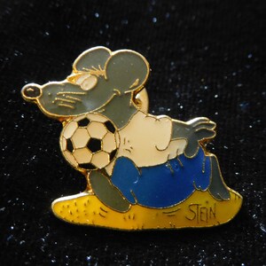 Puede incluir: Un pin de color dorado con un rat&oacute;n de dibujos animados que lleva un uniforme de f&uacute;tbol azul y blanco y sostiene un bal&oacute;n de f&uacute;tbol blanco y negro. El rat&oacute;n est&aacute; sentado en un campo amarillo con la palabra "STEIN" escrita en &eacute;l.