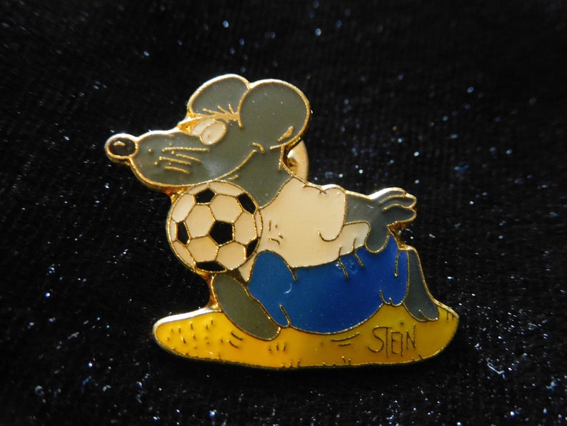 Puede incluir: Un pin de color dorado con un rat&oacute;n de dibujos animados que lleva un uniforme de f&uacute;tbol azul y blanco y sostiene un bal&oacute;n de f&uacute;tbol blanco y negro. El rat&oacute;n est&aacute; sentado en un campo amarillo con la palabra "STEIN" escrita en &eacute;l.