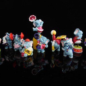 Könnte beinhalten: Ein Set mit 11 blauen Elefantenfiguren mit verschiedenen Zirkus-Accessoires. Die Elefanten tragen farbenfrohe Kostüme und halten Gegenstände wie eine Hantel, eine Trompete, einen Popcorn-Eimer und einen Ball.