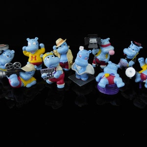 Può includere: Un set di dieci figurine di ippopotamo blu, ognuna con una posa e un accessorio diversi. Gli ippopotami indossano abiti diversi, tra cui una camicia gialla con una H, una camicia a righe rosse e bianche e una camicia a righe blu e bianche. Alcuni degli ippopotami tengono strumenti musicali, mentre altri tengono oggetti di scena come una bobina di pellicola e un microfono.