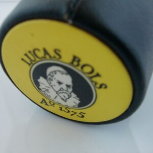 May include: Close-up of a yellow and black bottle cap with the text "Lucas Bols" and "A&deg; 1575" in black lettering. A black and white illustration of a man with a beard is in the center of the cap.