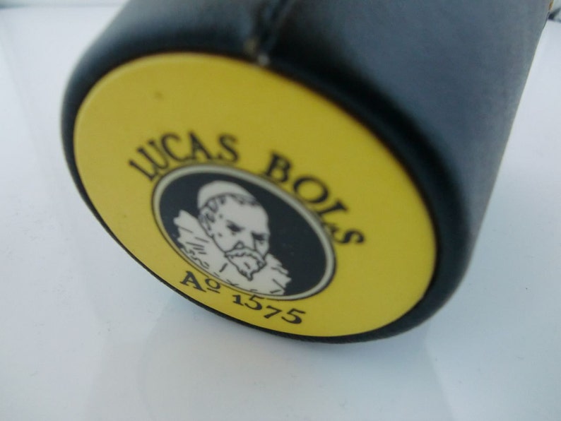 May include: Close-up of a yellow and black bottle cap with the text "Lucas Bols" and "A&deg; 1575" in black lettering. A black and white illustration of a man with a beard is in the center of the cap.