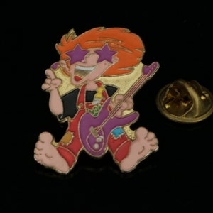 Könnte beinhalten: Eine goldfarbene Anstecknadel mit einem Cartoon-Charakter, der eine violette E-Gitarre spielt. Der Charakter hat rote Haare, ein schwarzes Hemd, eine rote Latzhose und trägt sternförmige Sonnenbrillen.