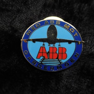 Könnte beinhalten: Eine blaue und goldene Anstecknadel mit einem weißen Flugzeug und dem Text "MED ABB MOT GARDERMOEN" in einem Kreis um das Flugzeug. Der Text "ABB" ist rot.