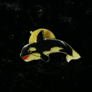 Könnte beinhalten: Eine schwarze und weiße Emaille-Anstecknadel mit einem Orca, oder Killerwal, mit rotem Mund. Die Nadel ist an einer goldfarbenen Rückseite befestigt.