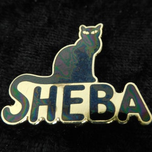 Könnte beinhalten: Eine goldfarbene Anstecknadel mit einer schwarzen Katzensilhouette und dem Wort "SHEBA" in schwarzen Buchstaben. Die Anstecknadel hat eine schillernde Oberfläche.
