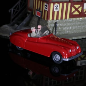 May include: A red toy car with two figures inside, driving on a black surface. The car has a white steering wheel and black wheels. The figures are wearing white shirts and black pants. The car is in front of a building with a sign that says "Riedlingen".