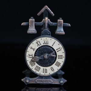 Könnte beinhalten: Ein Bleistiftspitzer im Vintage-Stil mit einem Zifferblatt und einem dekorativen Design. Der Spitzer ist aus Metall und hat ein schwarz-weißes Zifferblatt mit römischen Ziffern. Der Spitzer befindet sich auf einem schwarzen Hintergrund.