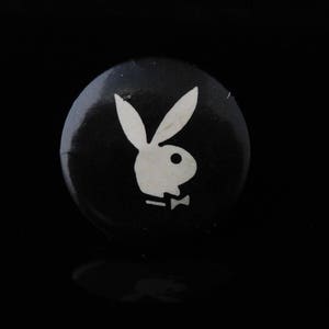 May include: A circular pin-back button featuring a black background with a white graphic of a rabbit head silhouette wearing a bow tie. The design is centered on the round surface of the button, which has a smooth, reflective finish.